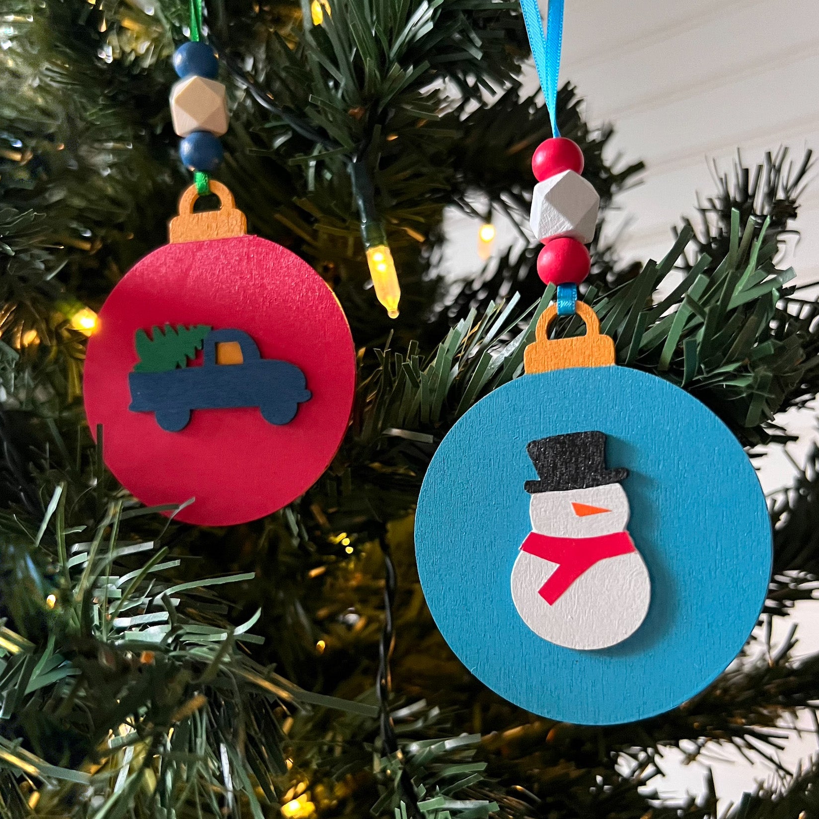 Image shows set of flat wood handmade ornaments. Each ornament features a holiday themed wood shape: a snowman and a truck bringing home the tree. Ornaments hang from ribbon and are decorated with three wood beads in matching colors.