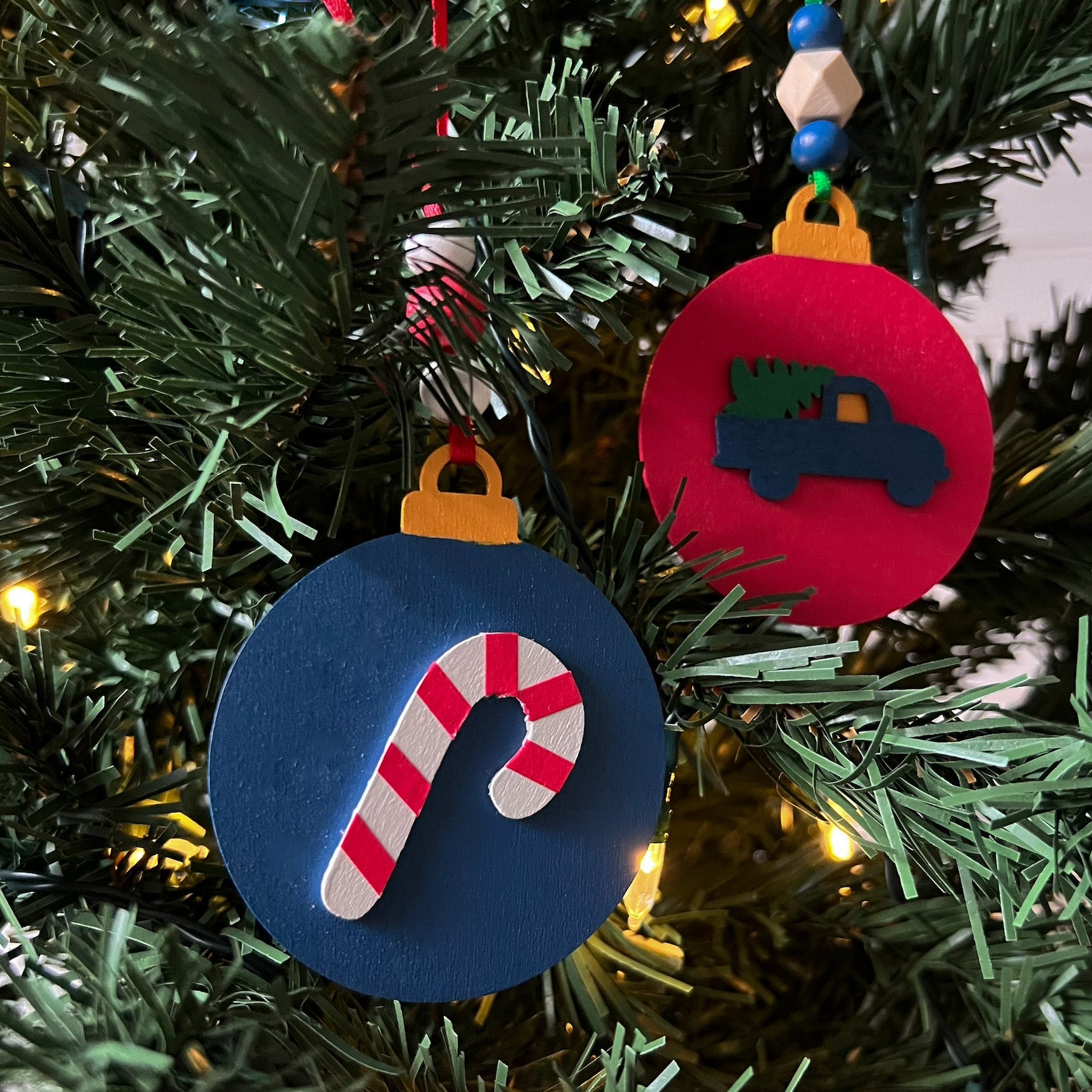 Image shows set of flat wood handmade ornaments. Each ornament features a holiday themed wood shape: a candy cane or a truck bringing home the tree. Ornaments hang from ribbon and are decorated with three wood beads in matching colors.
