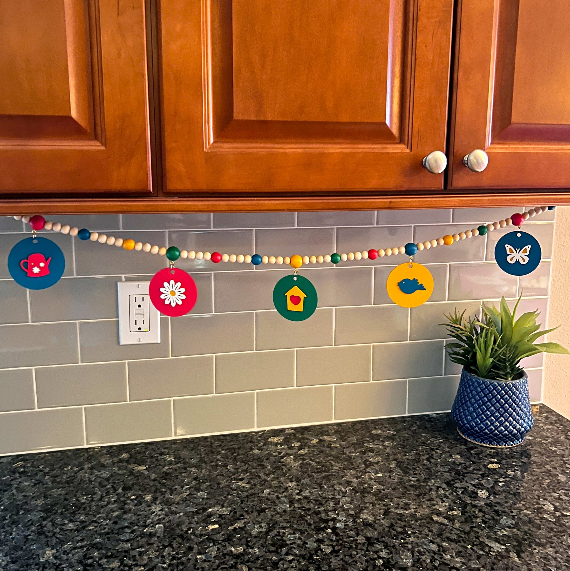 In the Garden Handmade Garland. The garland features wood circles a garden themed shape – a watering can, flower, birdhouse, bluebird, or butterfly. Shapes hang from wood beads. Spring or summer garden seasonal decoration or gift.