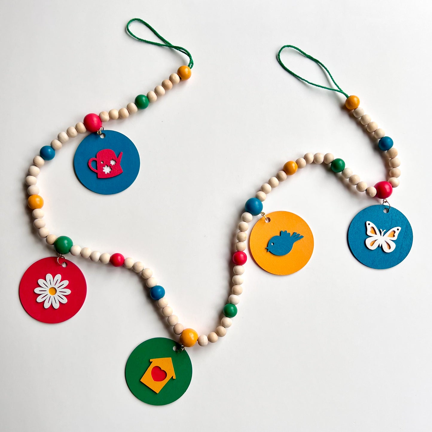 In the Garden Handmade Garland. The garland features wood circles a garden themed shape – a watering can, flower, birdhouse, bluebird, or butterfly. Shapes hang from wood beads. Spring or summer garden seasonal decoration or gift.