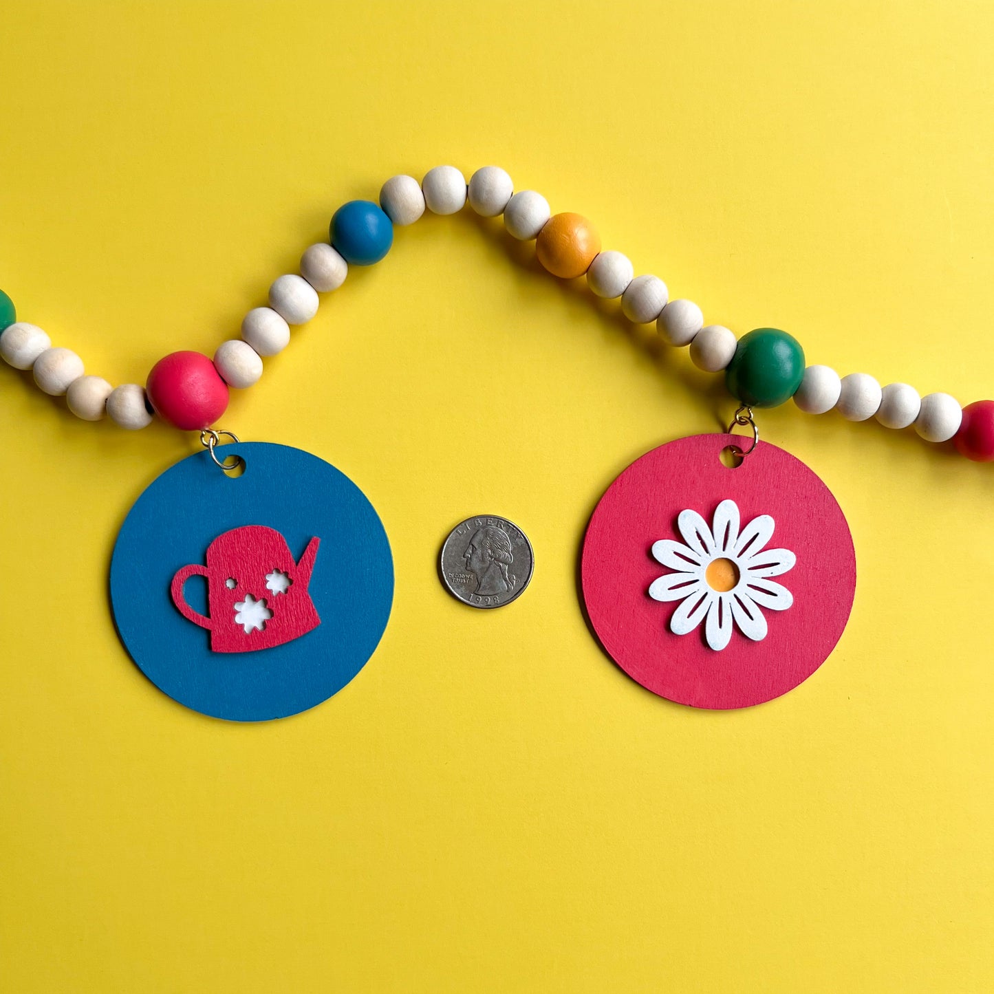 In the Garden Handmade Garland. The garland features wood circles a garden themed shape – a watering can, flower, birdhouse, bluebird, or butterfly. Shapes hang from wood beads. Spring or summer garden seasonal decoration or gift.