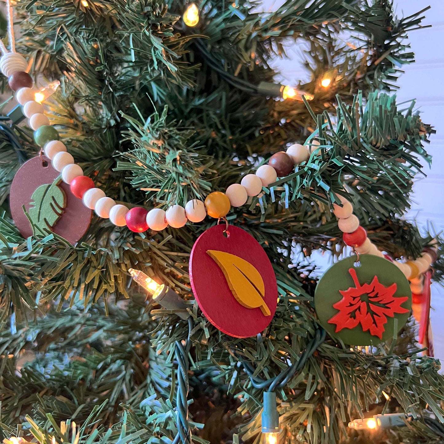 Image shows a wood bead garland with 2" wood circles that feature various colored fall leaves. Circles hang from painted beads with natural wood beads interspersed. Beads are strung on 2mm orange macrame cord with 3" loops on each end.