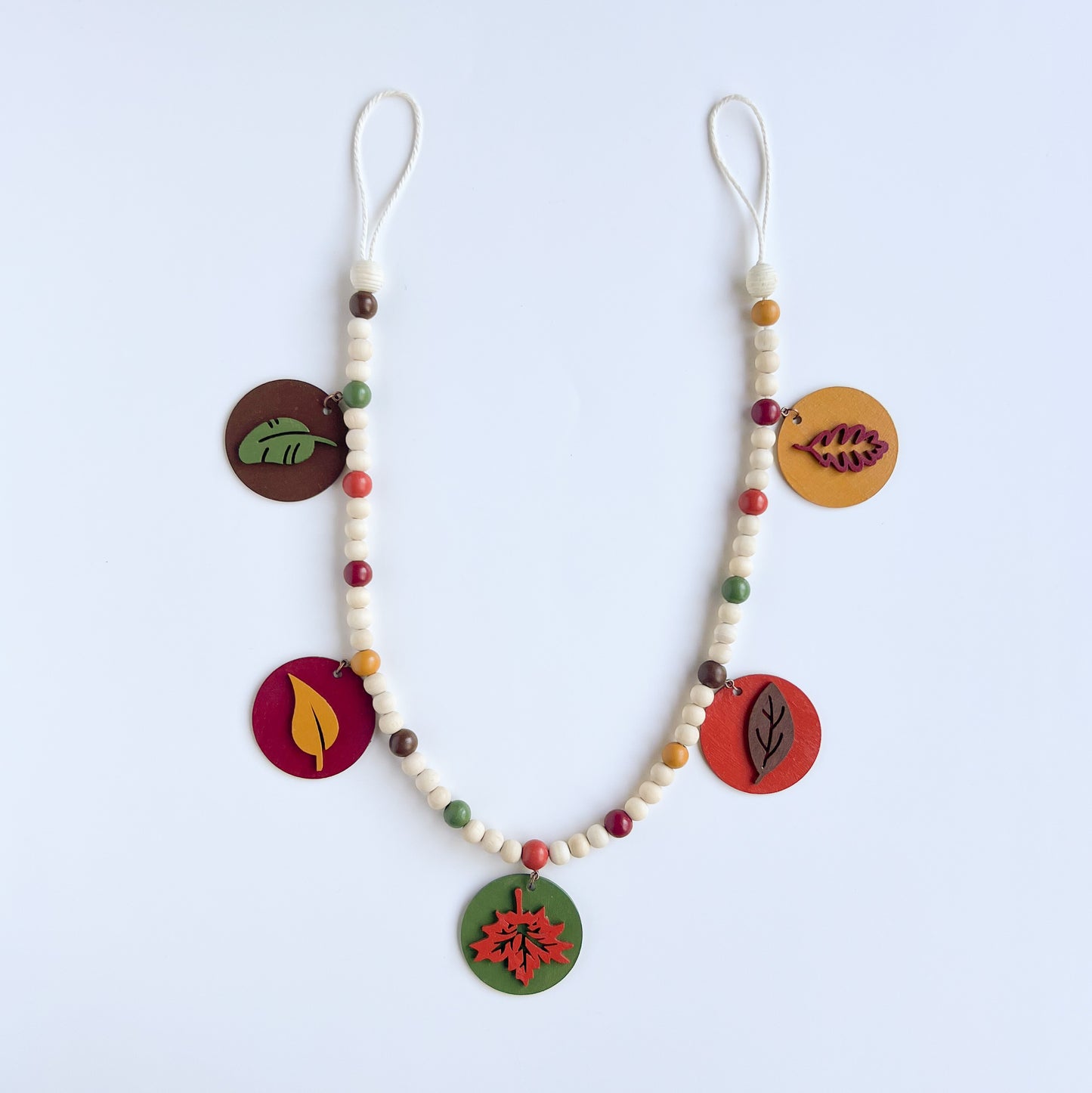Image shows a wood bead garland with 2" wood circles that feature various colored fall leaves. Circles hang from painted beads with natural wood beads interspersed. Beads are strung on 2mm orange macrame cord with 3" loops on each end.