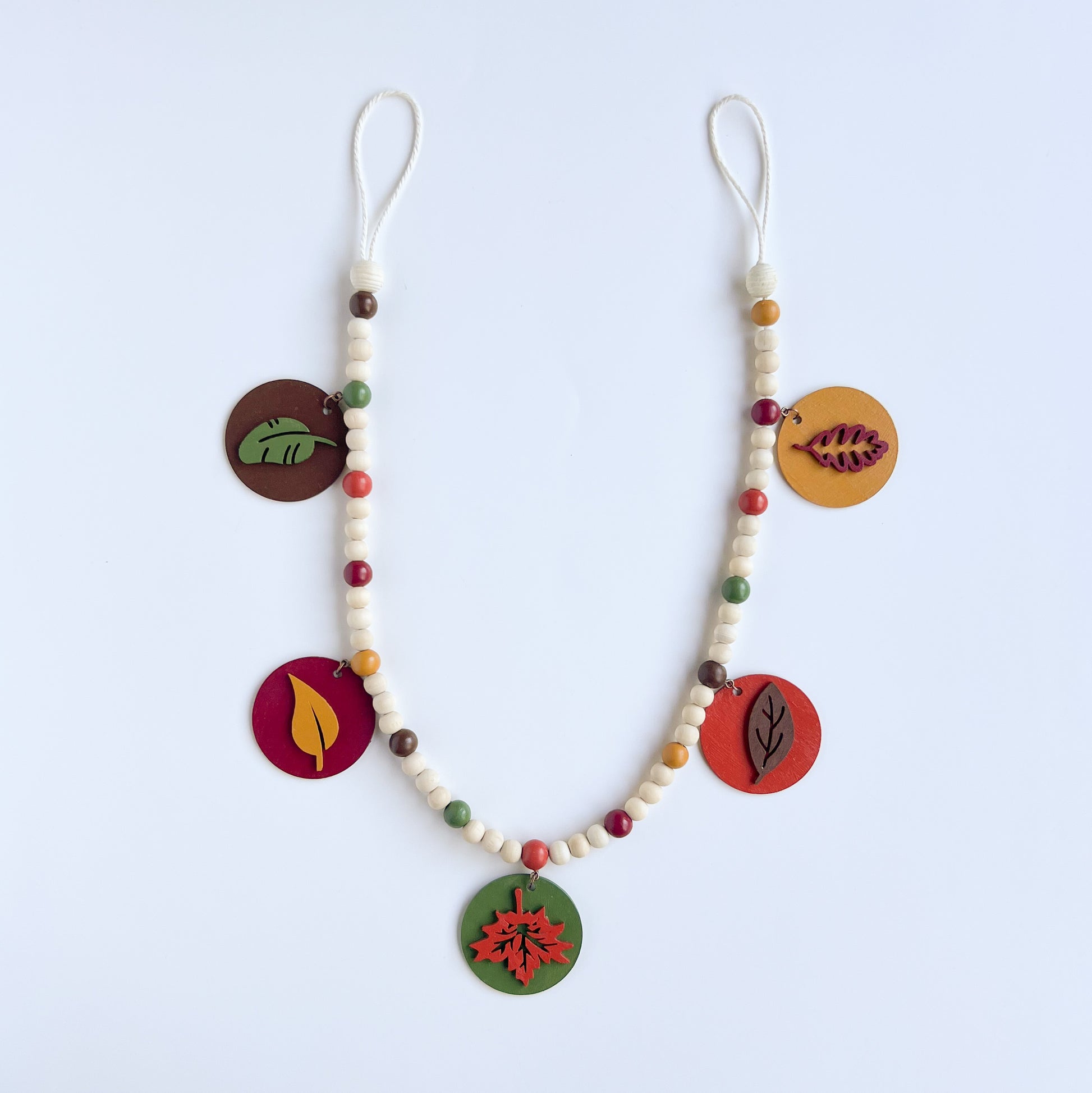 Image shows a wood bead garland with 2" wood circles that feature various colored fall leaves. Circles hang from painted beads with natural wood beads interspersed. Beads are strung on 2mm orange macrame cord with 3" loops on each end.
