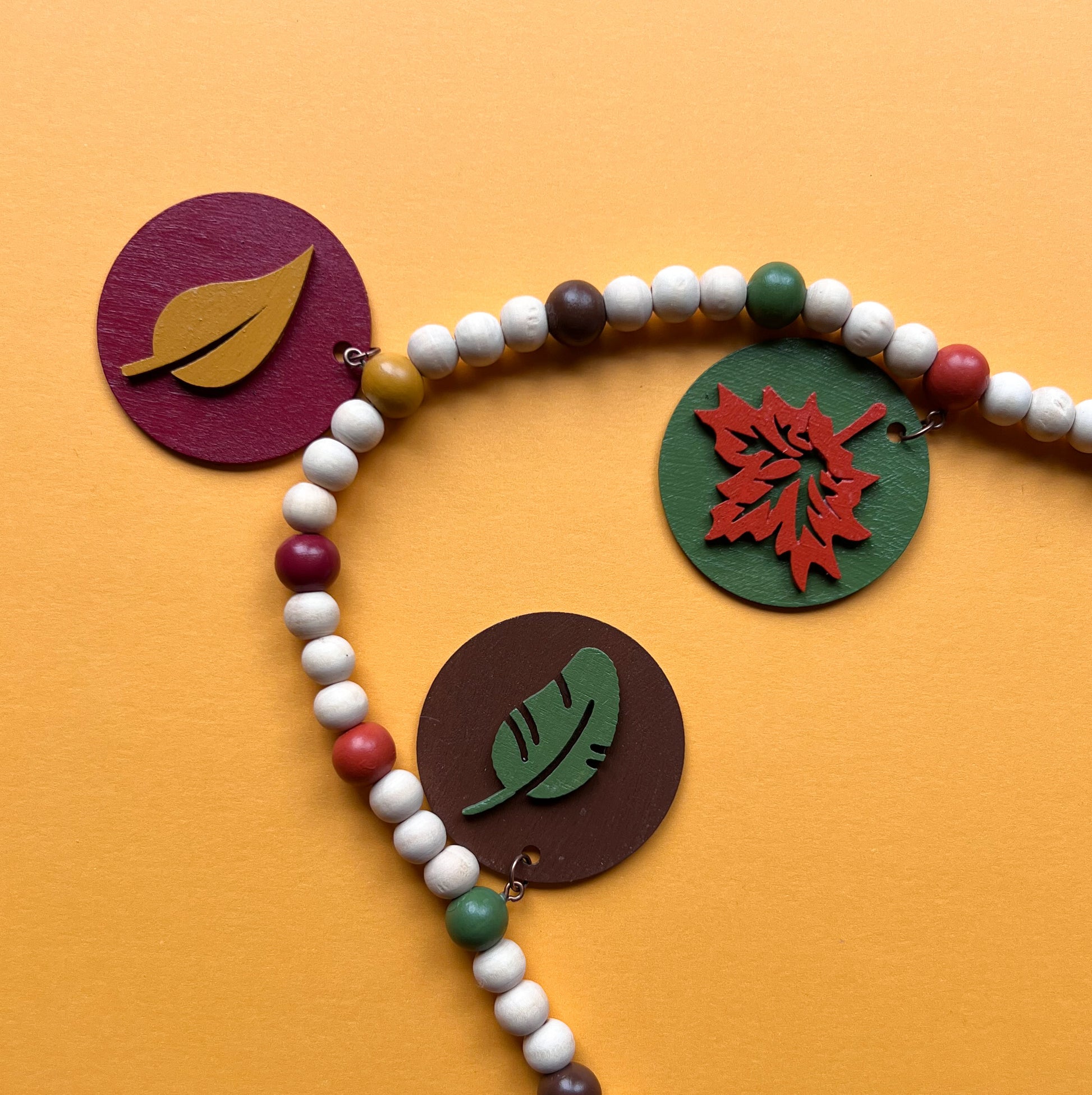 Image shows a wood bead garland with 2" wood circles that feature various colored fall leaves. Circles hang from painted beads with natural wood beads interspersed. Beads are strung on 2mm orange macrame cord with 3" loops on each end.