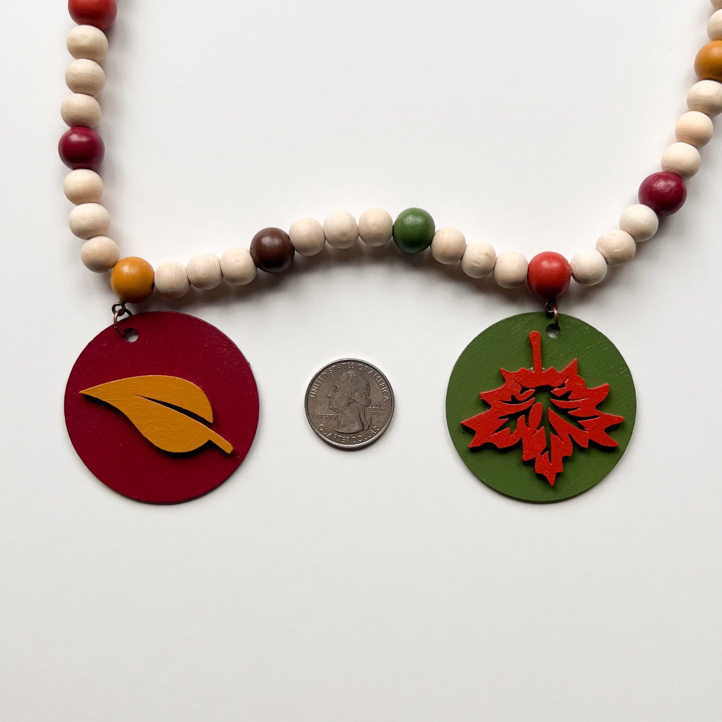 Image shows a wood bead garland with 2" wood circles that feature various colored fall leaves. Circles hang from painted beads with natural wood beads interspersed. Beads are strung on 2mm orange macrame cord with 3" loops on each end.