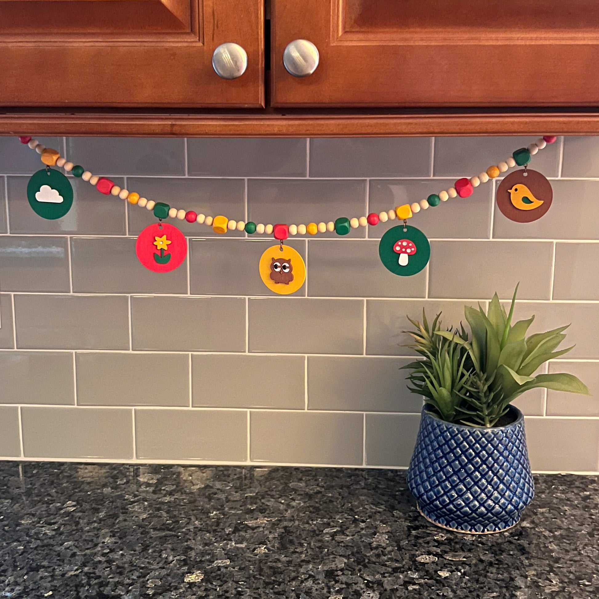Image shows handmade garland with wood circles featuring nature themed shapes. Wood circles hang from painted square beads and are strung with other painted and natural round wood beads. Includes 3-inch loops on either end.
