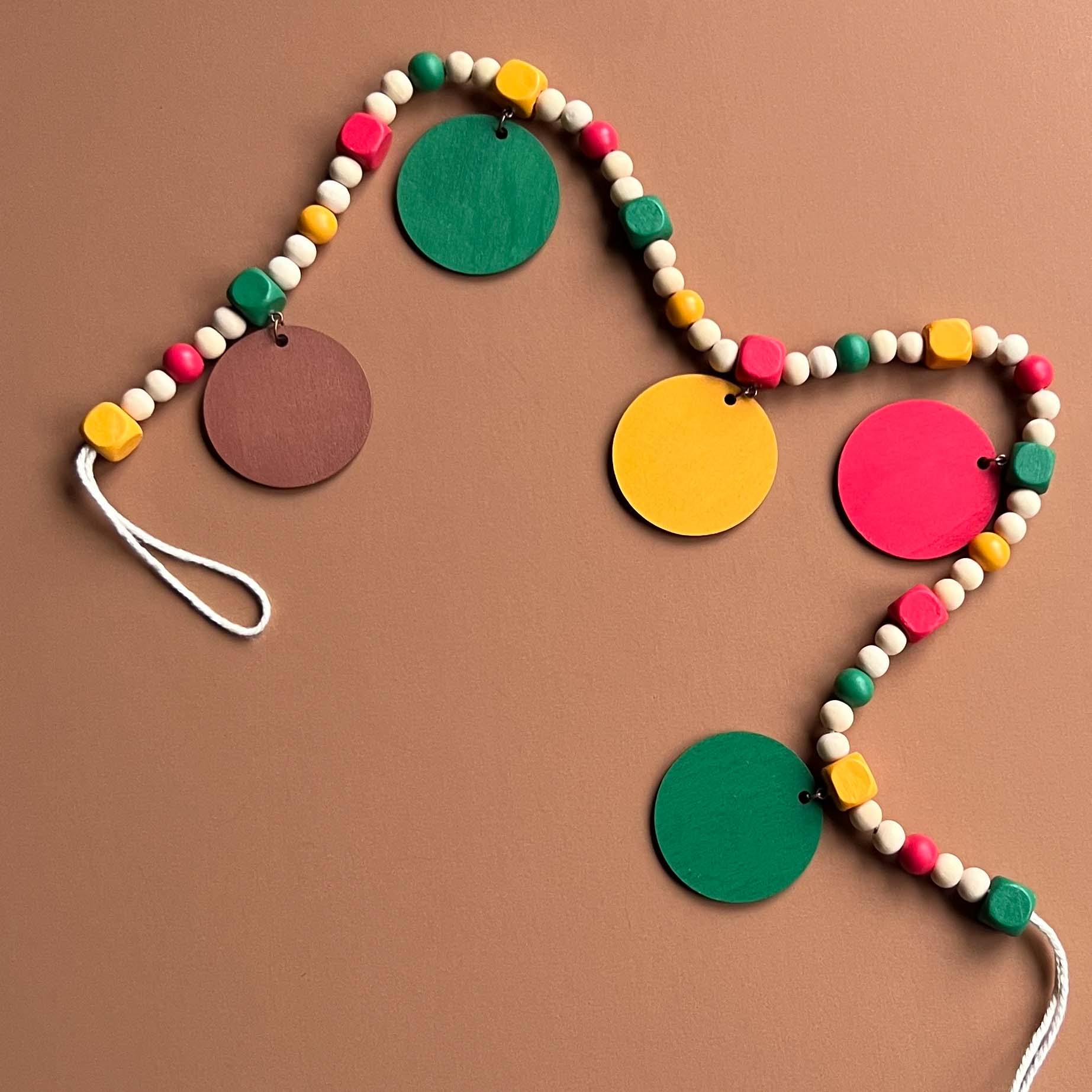 Image shows handmade garland with wood circles featuring nature themed shapes. Wood circles hang from painted square beads and are strung with other painted and natural round wood beads. Includes 3-inch loops on either end.