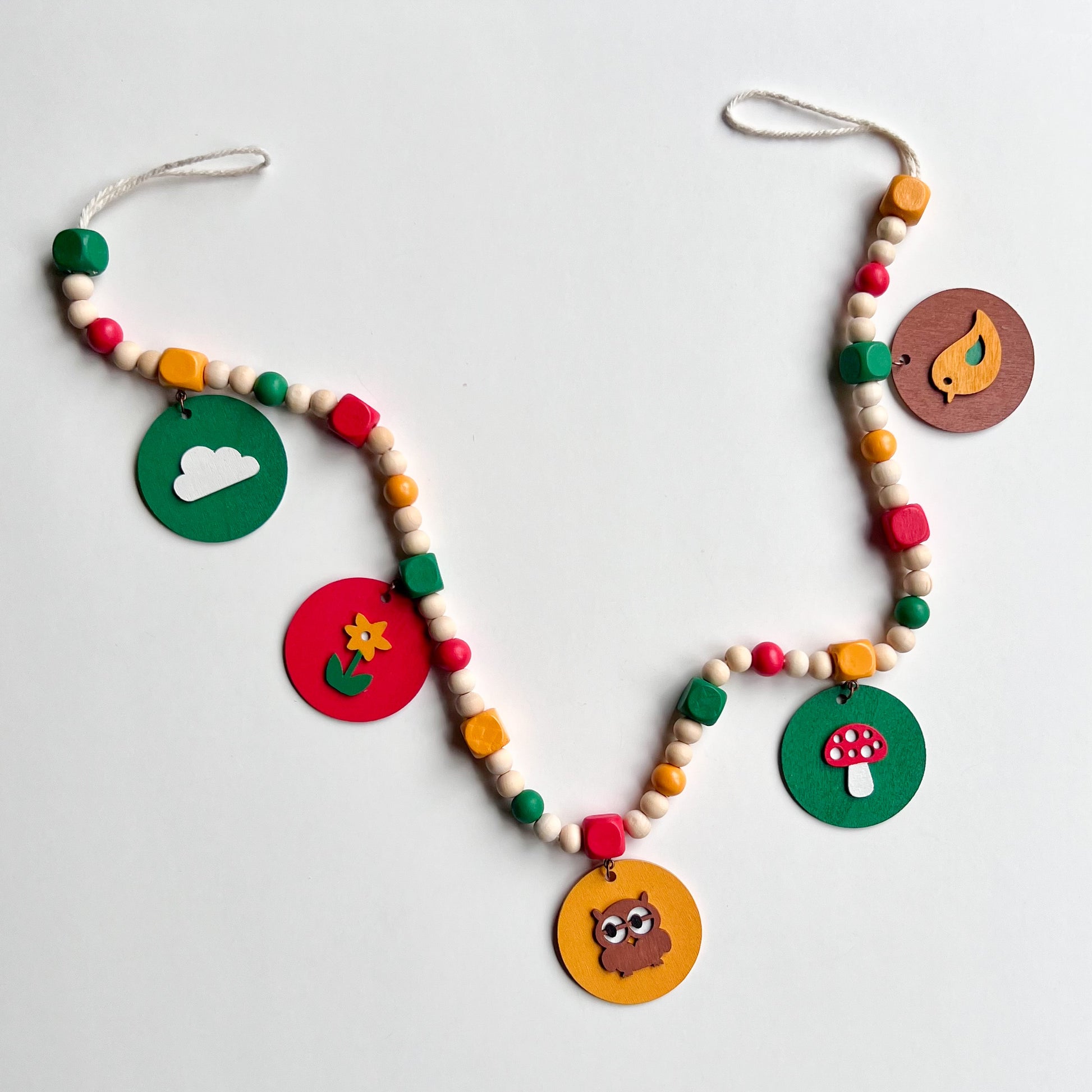 Image shows handmade garland with wood circles featuring nature themed shapes. Wood circles hang from painted square beads and are strung with other painted and natural round wood beads. Includes 3-inch loops on either end.