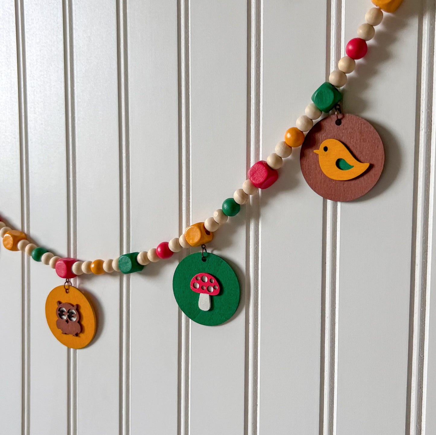 Image shows handmade garland with wood circles featuring nature themed shapes. Wood circles hang from painted square beads and are strung with other painted and natural round wood beads. Includes 3-inch loops on either end.
