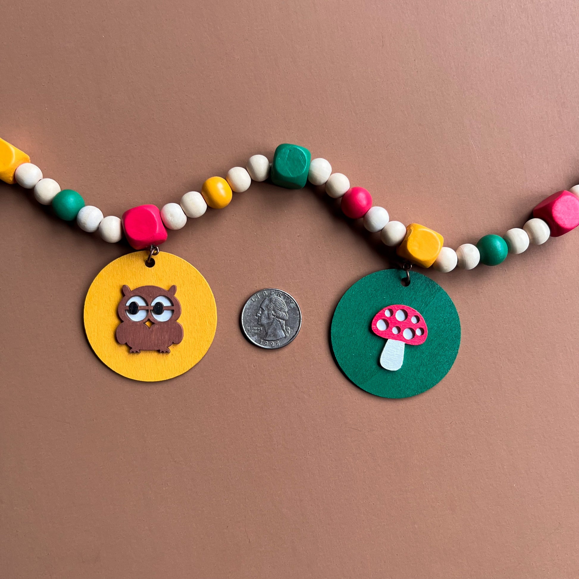 Image shows handmade garland with wood circles featuring nature themed shapes. Wood circles hang from painted square beads and are strung with other painted and natural round wood beads. Includes 3-inch loops on either end.