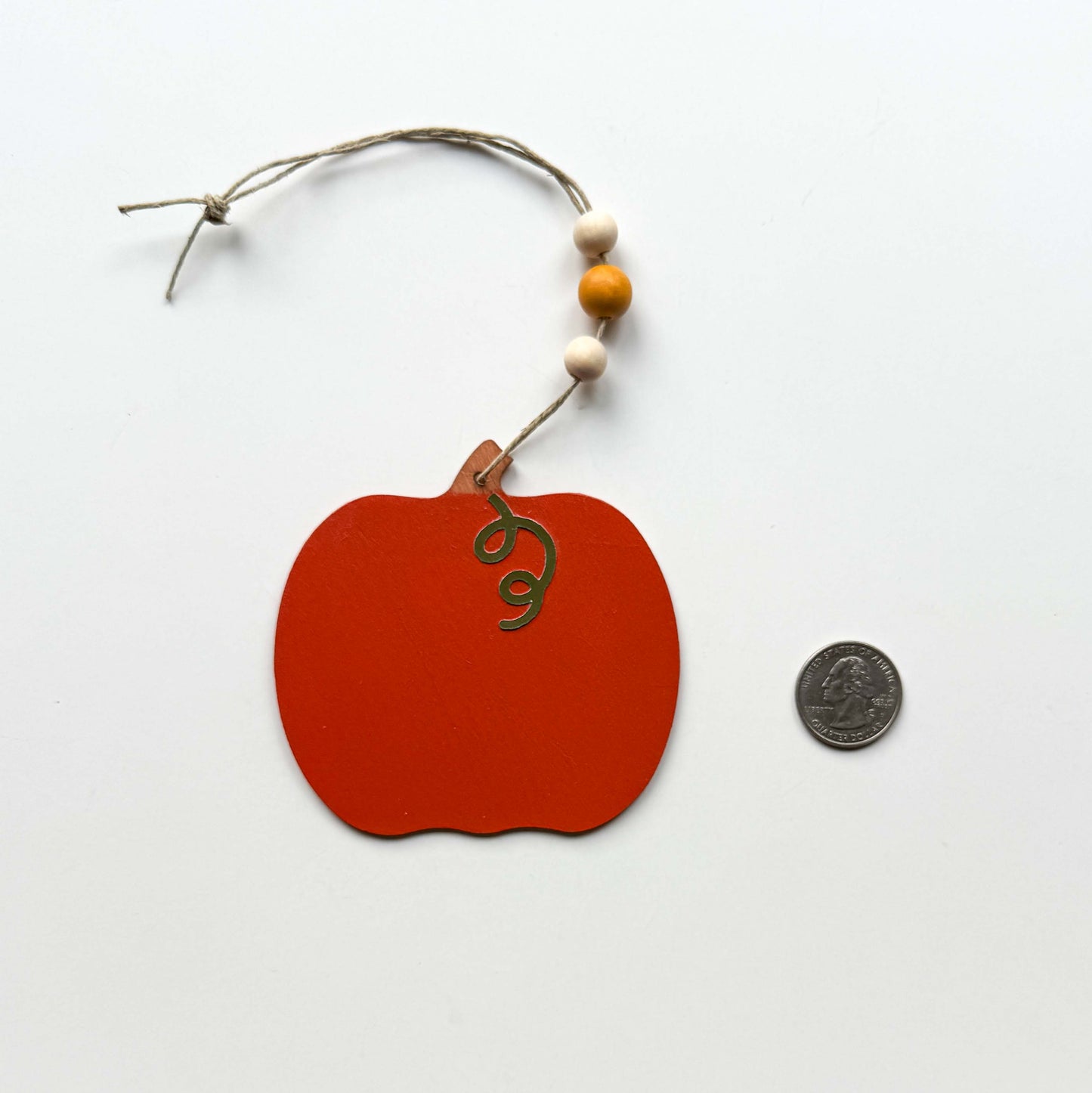 Image of a wood pumpkin ornament painted a rust orange. The ornament hangs from twine and feature three wood beads. A quarter is placed next to the ornament for a size comparison.
