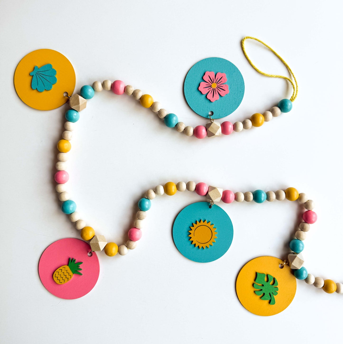 Summer themed wood bead garland with natural, pink, blue, and yellow beads. 5 wood circles hang from geometric beads and feature tropical themed shapes. Garland is placed on a white background.