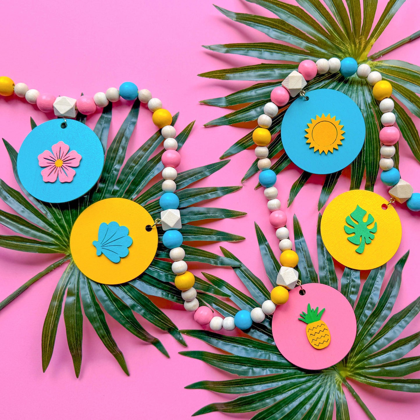 Summer themed wood bead garland with natural, pink, blue, and yellow beads. 5 wood circles hang from geometric beads and feature tropical themed shapes. Garland is placed on a pink background with palm leaves.