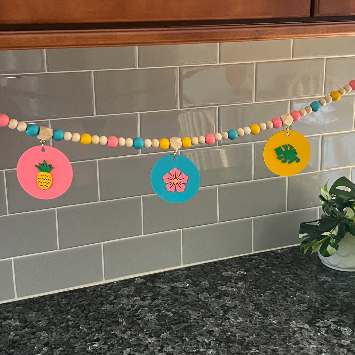 Back of summer themed wood bead garland with natural, pink, blue, and yellow beads. 5 wood circles hang from geometric beads and feature tropical themed shapes. Garland is hanging under cabinets in a kitchen.