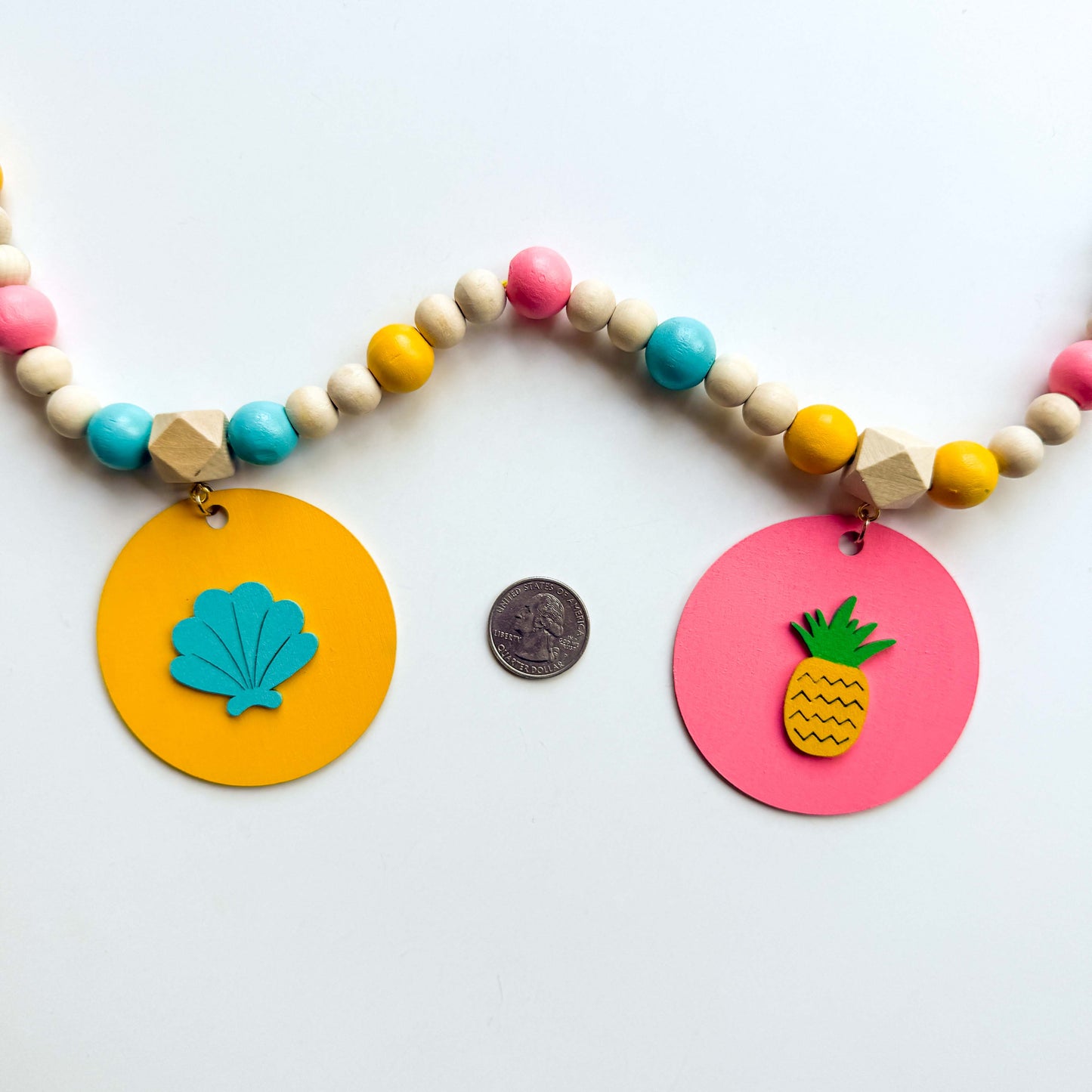 Back of summer themed wood bead garland with natural, pink, blue, and yellow beads. 5 wood circles hang from geometric beads and feature tropical themed shapes. Circles with shell and pineapple are shown with a quarter in between them to show size.