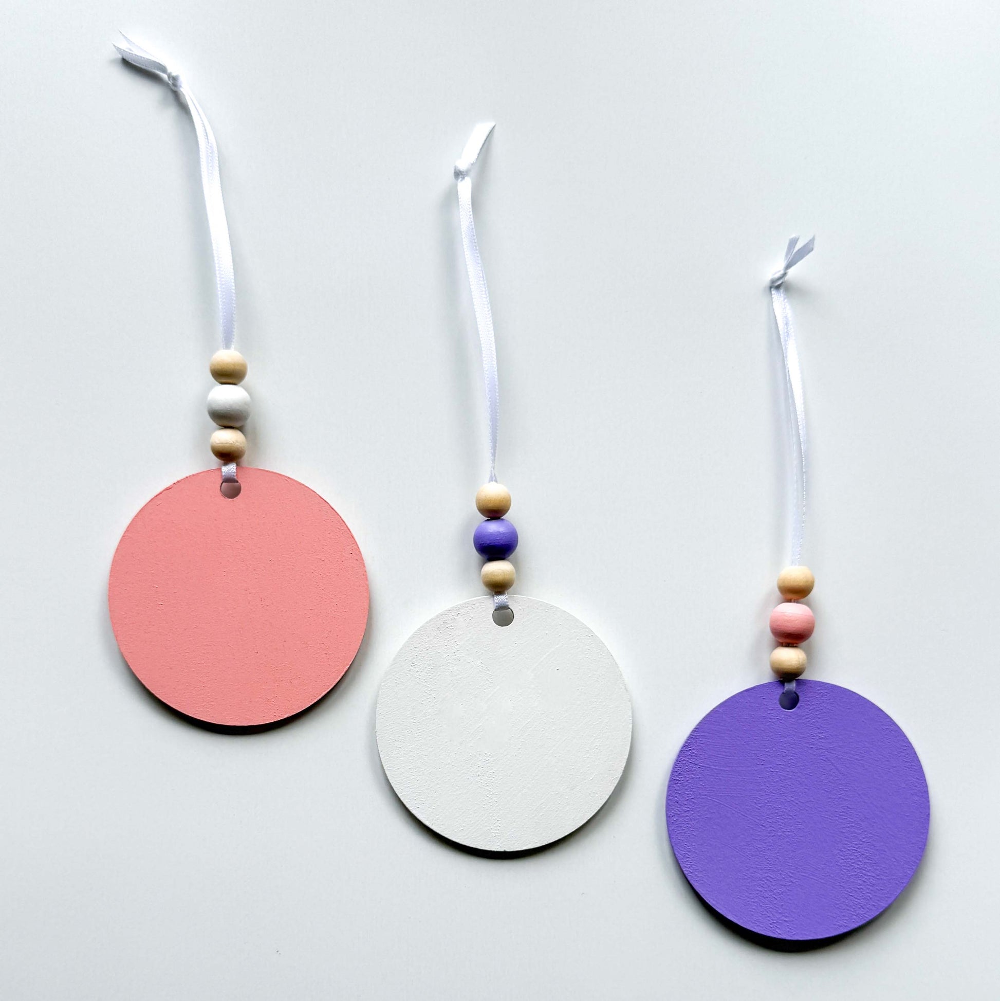 Back of three spring themed ornaments in white, lavender, and light pink. Ornaments each feature a different spring shape - a flower, a birdhouse, or a butterfly. Each ornament hangs from a white ribbon with three wood beads as an accent.