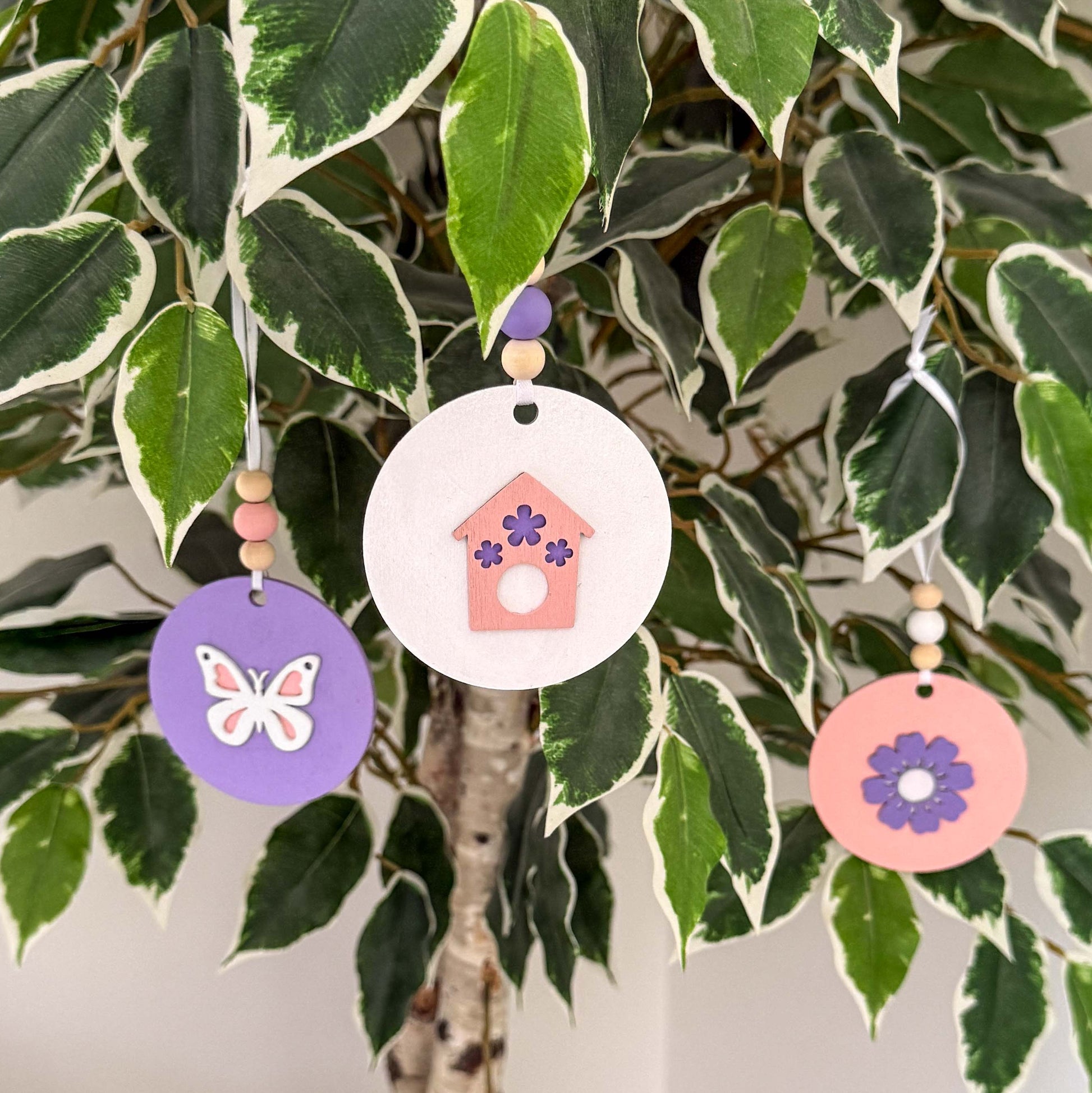 Three spring themed ornaments in white, lavender, and light pink. Ornaments each feature a different spring shape - a flower, a birdhouse, or a butterfly. Each ornament hangs from a white ribbon with three wood beads as an accent. Ornaments hang from a indoor tree as an accent.