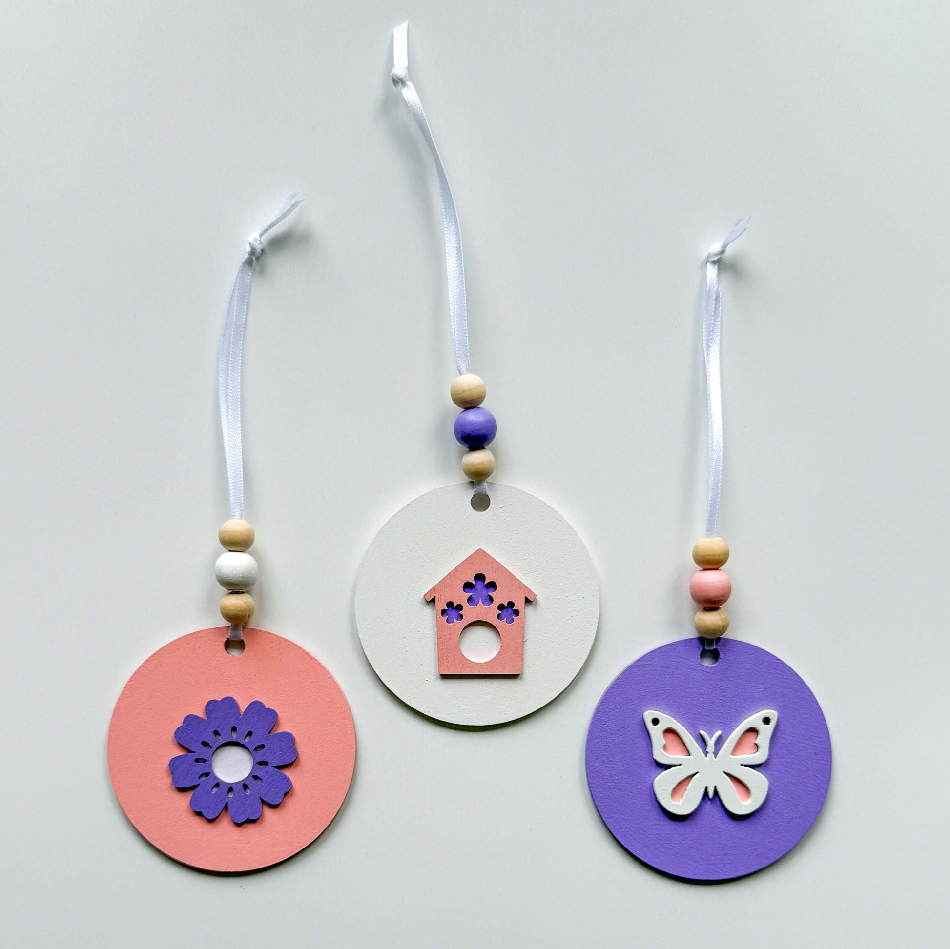 Three spring themed ornaments in white, lavender, and light pink. Ornaments each feature a different spring shape - a flower, a birdhouse, or a butterfly. Each ornament hangs from a white ribbon with three wood beads as an accent.