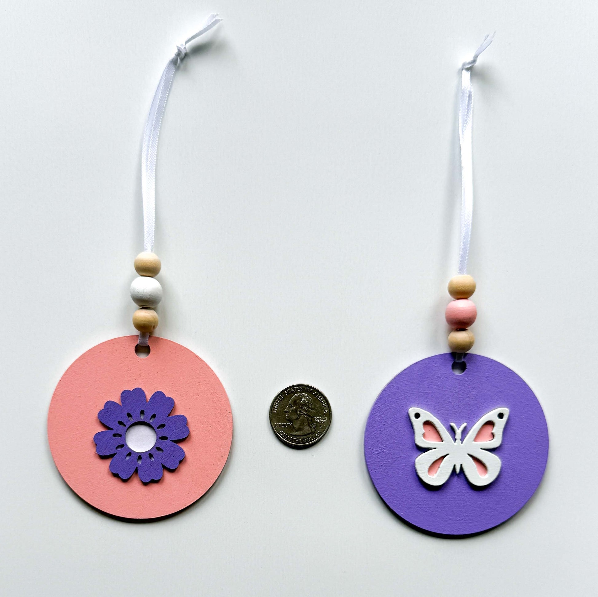 Back of three spring themed ornaments in white, lavender, and light pink. Ornaments each feature a different spring shape - a flower, a birdhouse, or a butterfly. Each ornament hangs from a white ribbon with three wood beads as an accent. Image features a pink ornament and a lavender ornament next to a quarter for size comparison.