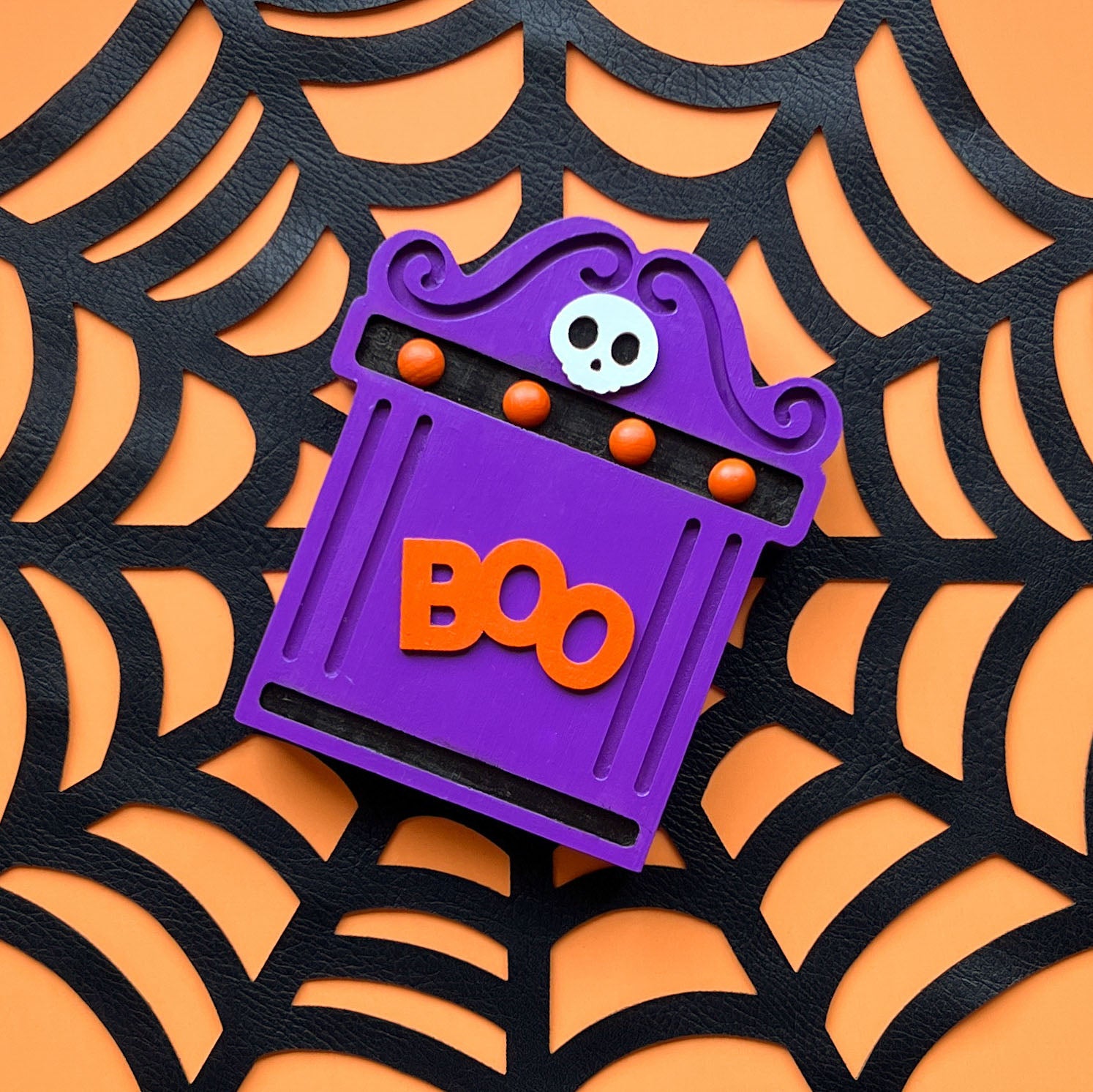 Image of a purple wood tombstone shelf sitter with orange and black painted accents and a small white wood skull. Shelf sitter is shown on a large black cobweb.