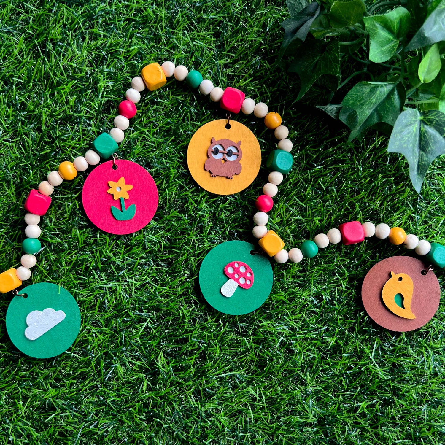 Image of a small wood bead garland with circle elements that hang from square beads. These circles feature a cloud, flower, owl, toadstool, or bird. Garland is placed on a faux grass background.