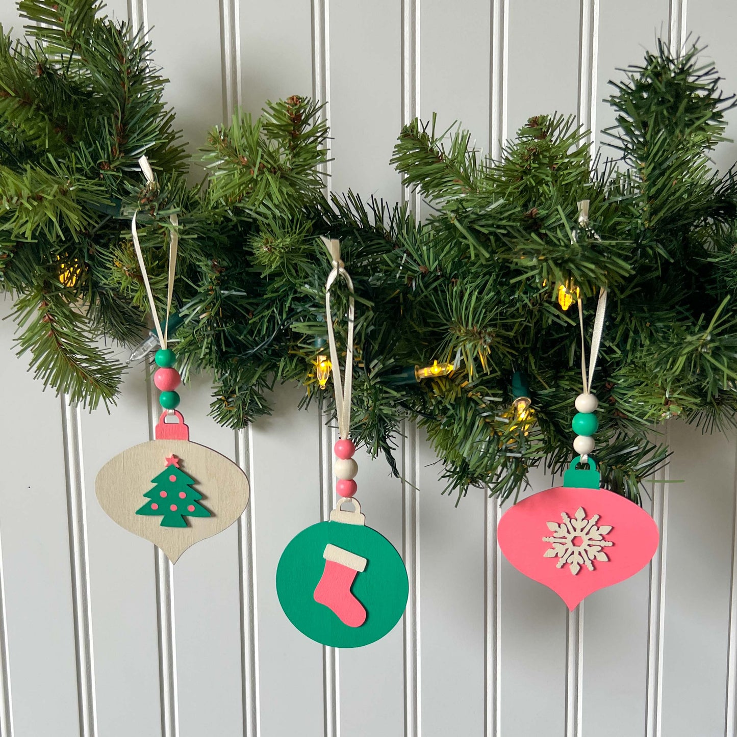 Retro themed wood Christmas ornaments painted bright pink, teal, and antique white. Ornaments feature wood shapes including a snowflake, a stocking, and a decorated tree. Each ornament hangs from a cream ribbon with three painted wood beads.