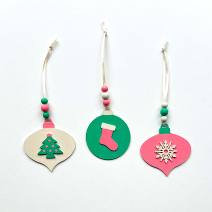 Retro themed wood Christmas ornaments painted bright pink, teal, and antique white. Ornaments feature wood shapes including a snowflake, a stocking, and a decorated tree. Each ornament hangs from a cream ribbon with three painted wood beads.