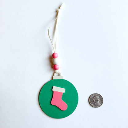 Retro themed wood Christmas ornaments painted bright pink, teal, and antique white. Ornaments feature wood shapes including a snowflake, a stocking, and a decorated tree. Each ornament hangs from a cream ribbon with three painted wood beads.