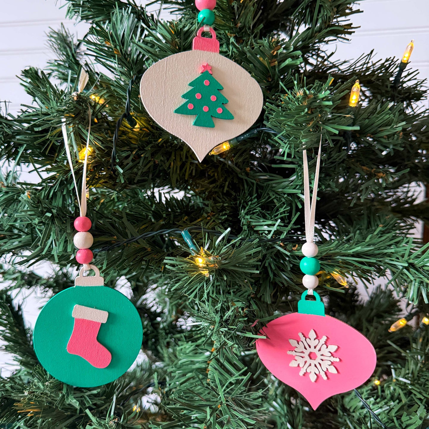 Retro themed wood Christmas ornaments painted bright pink, teal, and antique white. Ornaments feature wood shapes including a snowflake, a stocking, and a decorated tree. Each ornament hangs from a cream ribbon with three painted wood beads.