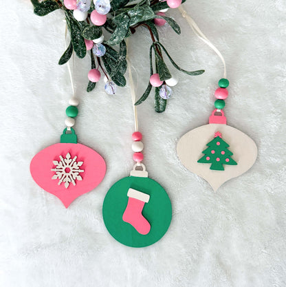 Retro themed wood Christmas ornaments painted bright pink, teal, and antique white. Ornaments feature wood shapes including a snowflake, a stocking, and a decorated tree. Each ornament hangs from a cream ribbon with three painted wood beads.