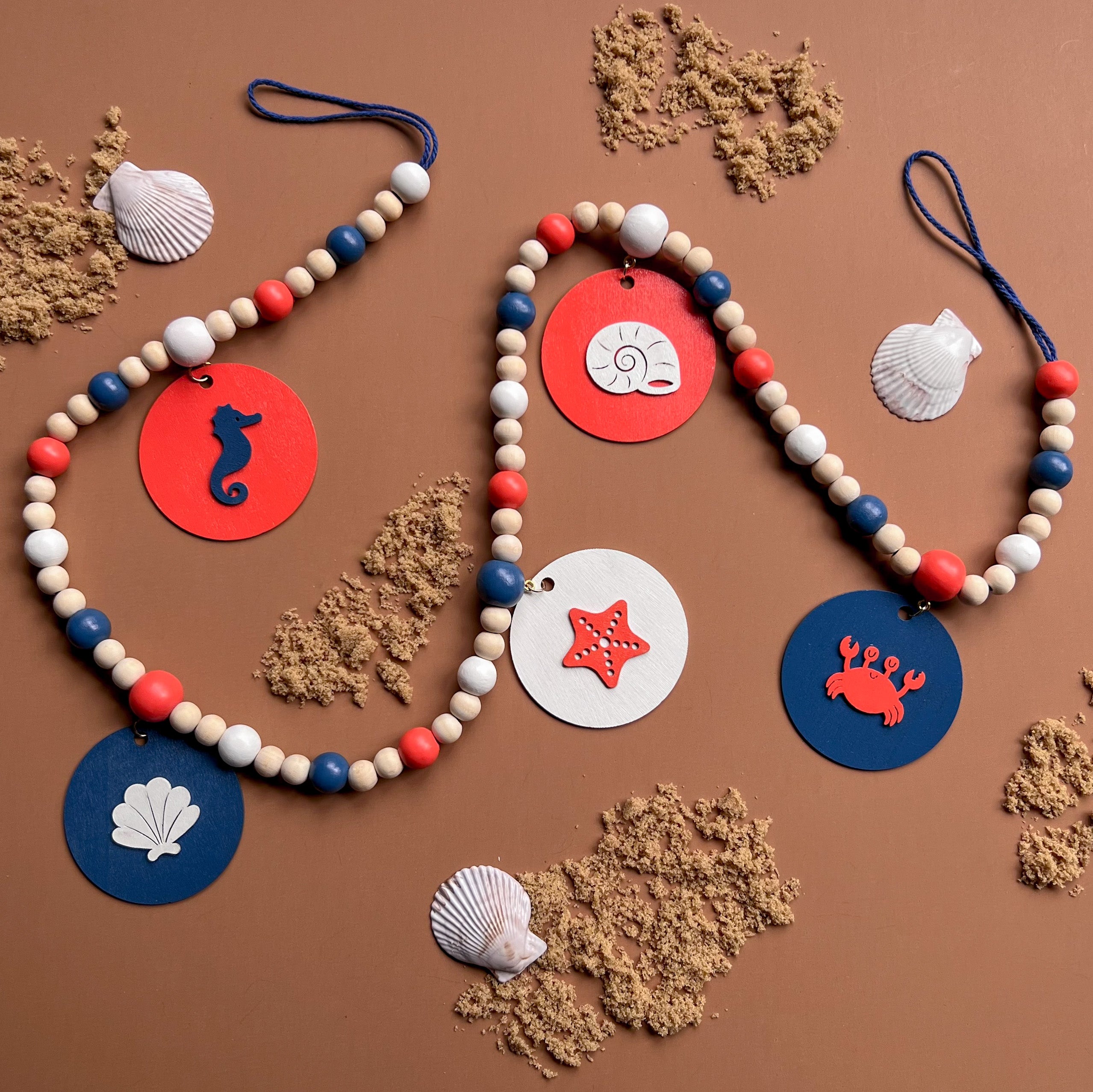 At the Seashore Handmade Garland. Features beachy wood shapes: seahorse, shells, starfish, and a crab. Shapes hang from painted wood beads. Summer beach seaside decor.