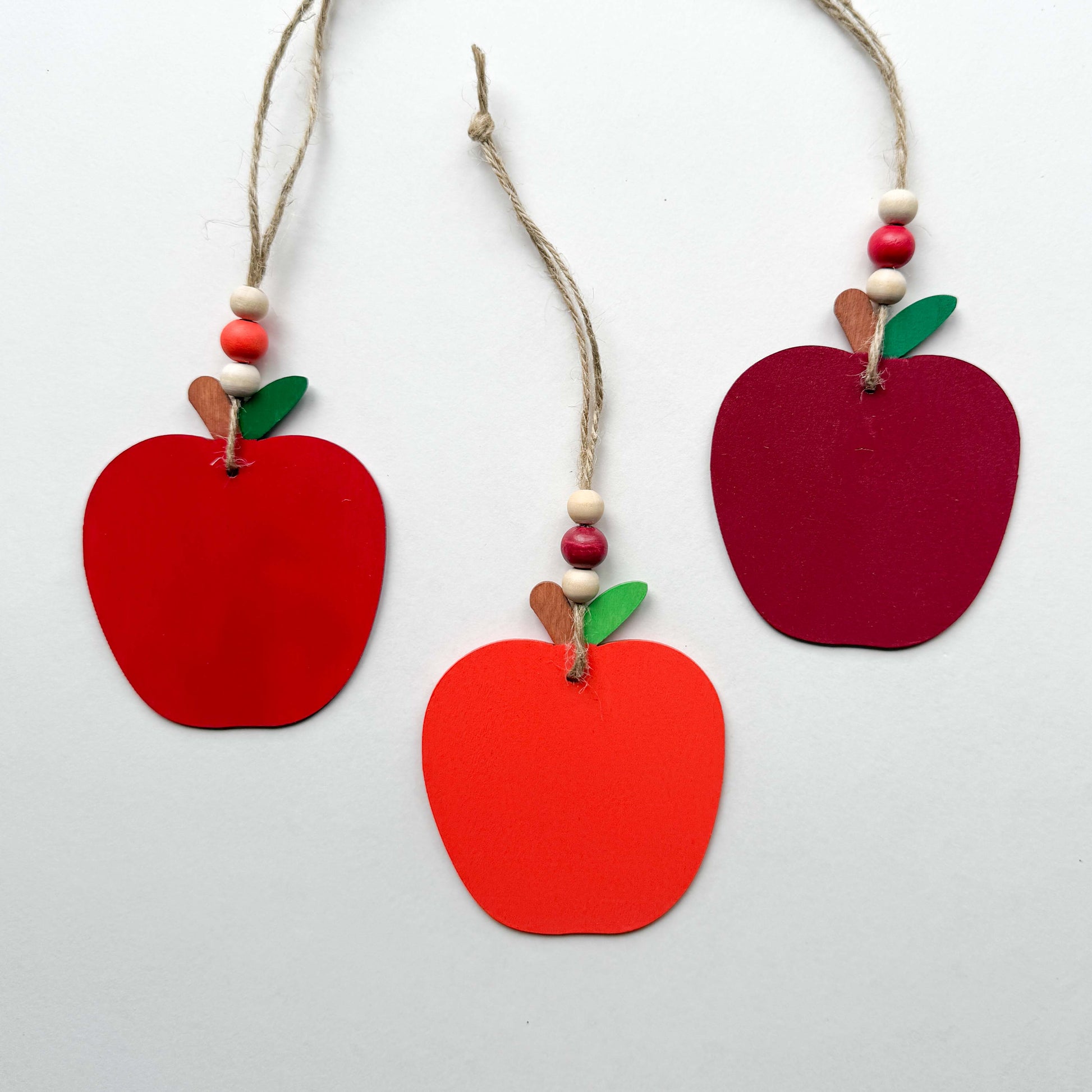Three wood apple ornaments, each painted in a different shade of red. Ornaments hang from jute twine and feature three wood beads on each.