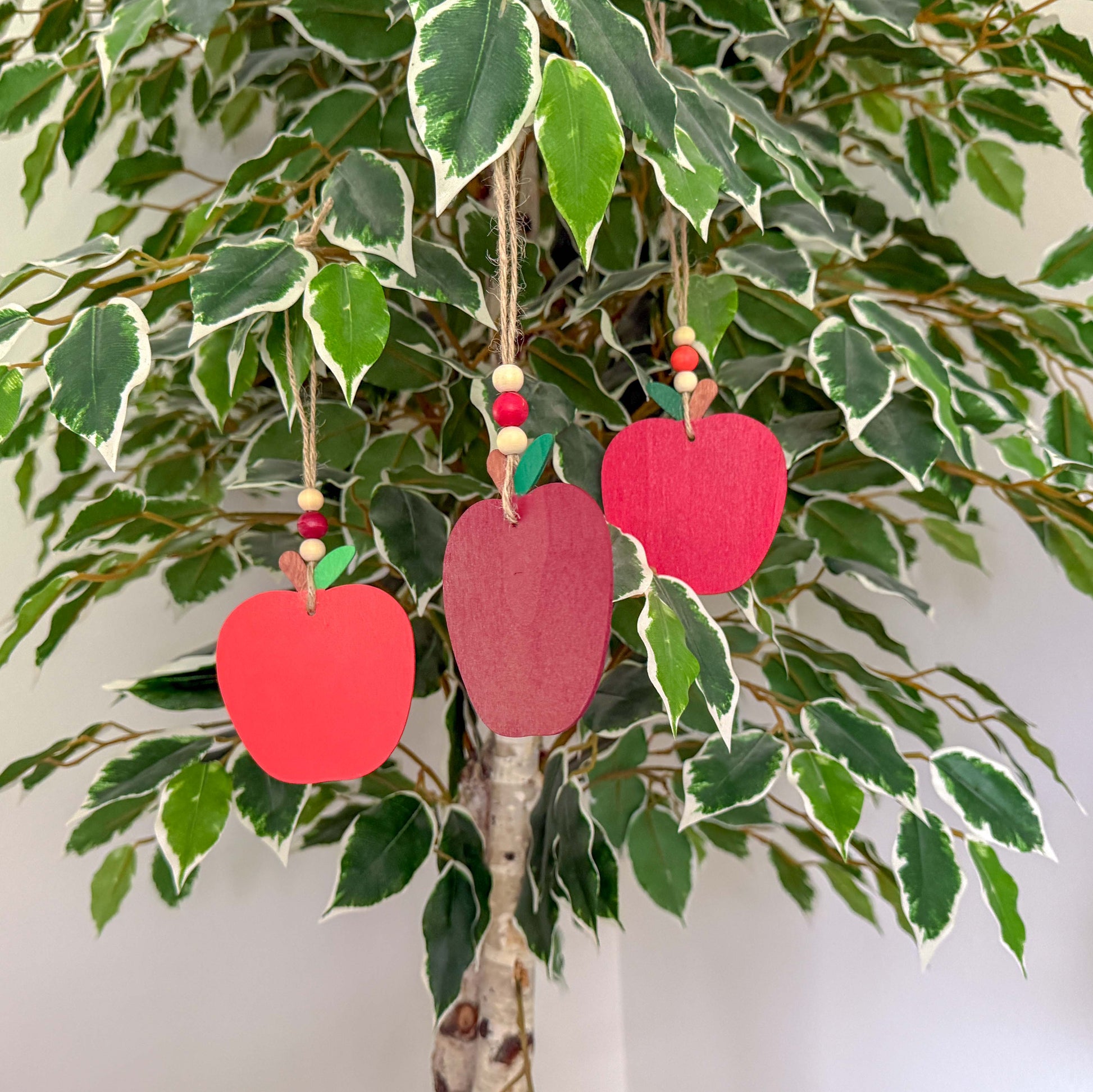 Three wood apple ornaments, each painted in a different shade of red. Ornaments hang from jute twine and feature three wood beads on each.