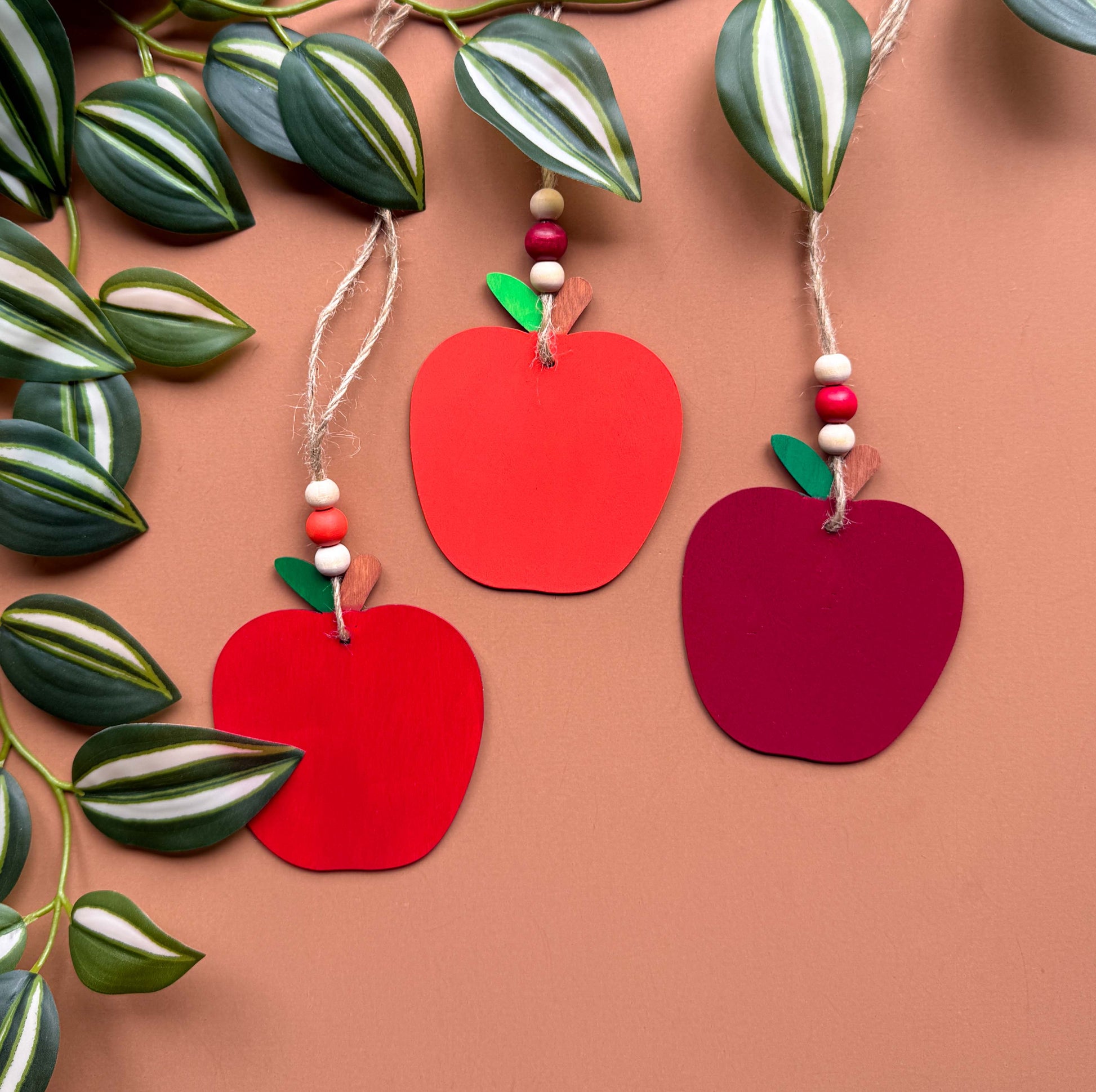 Three wood apple ornaments, each painted in a different shade of red. Ornaments hang from jute twine and feature three wood beads on each.
