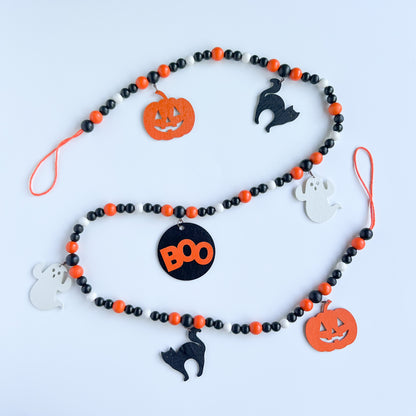 Boo to You Handmade Garland. The garland features seven Halloween themed shapes – jack-o-lanterns, ghosts, black cats, and a wood boo element in the center. Shapes hang from painted wood beads. Halloween fall seasonal decoration or gift.