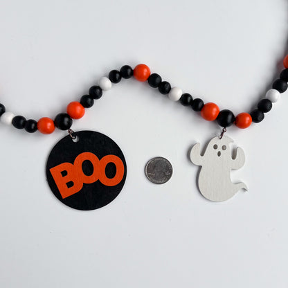 Boo to You Handmade Garland. The garland features seven Halloween themed shapes – jack-o-lanterns, ghosts, black cats, and a wood boo element in the center. Shapes hang from painted wood beads. Halloween fall seasonal decoration or gift.