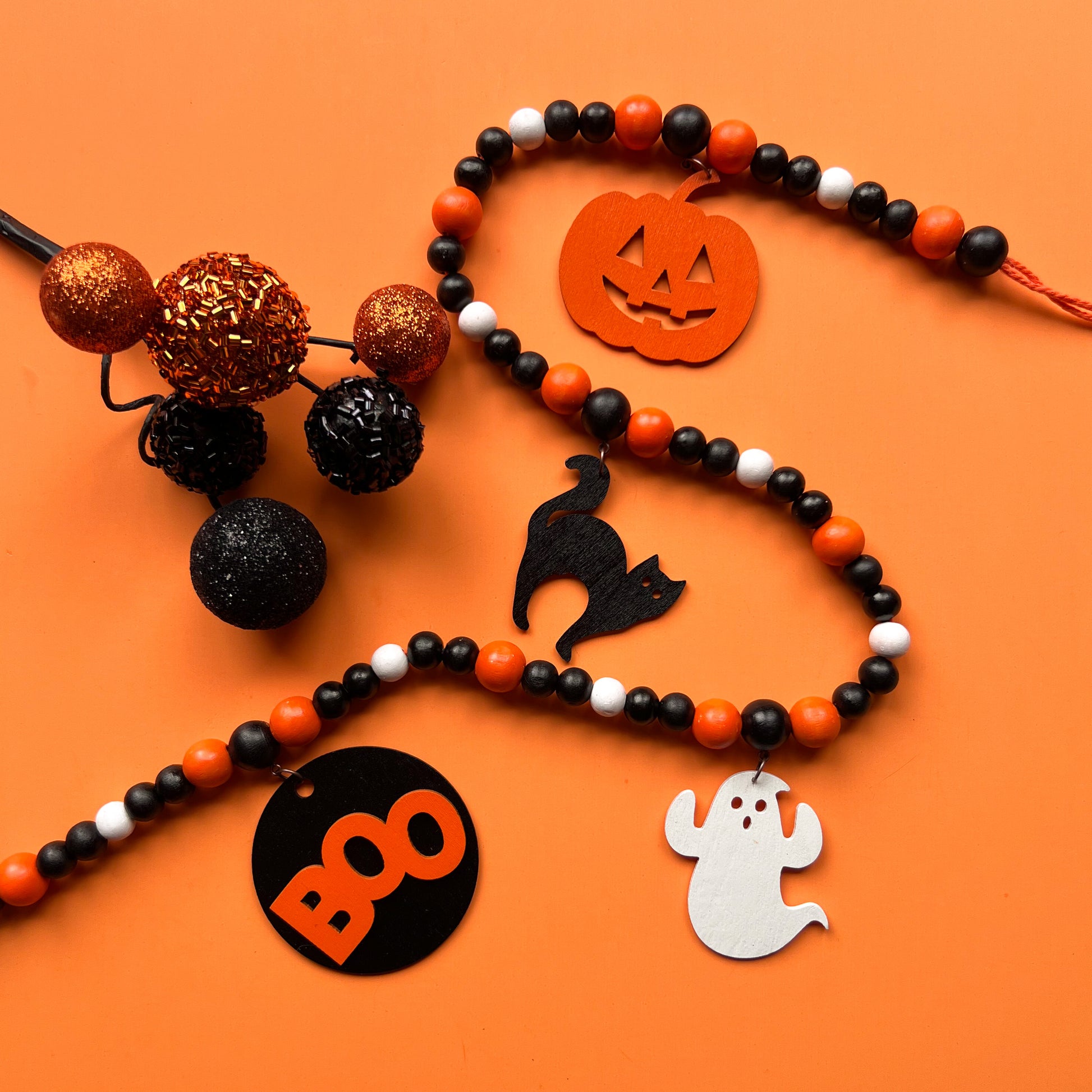 Boo to You Handmade Garland. The garland features seven Halloween themed shapes – jack-o-lanterns, ghosts, black cats, and a wood boo element in the center. Shapes hang from painted wood beads. Halloween fall seasonal decoration or gift.