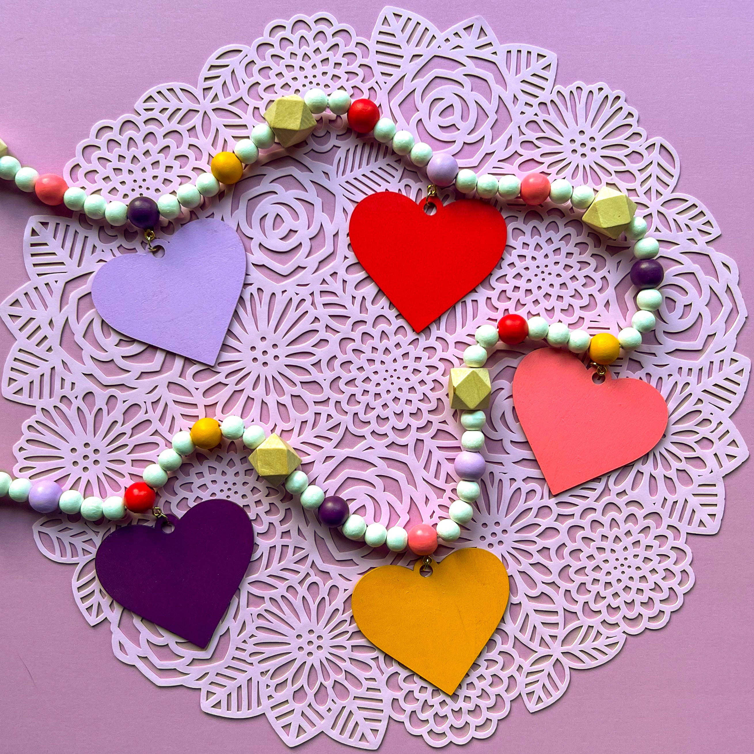 Images shows a garland with wood hearts in various colors – lavender, plum, golden yellow, bright pink, and pinkish-red. Hearts hang from natural geometric wood beads with a gold jump ring and eye screw. Those beads are strung with beads in corresponding colors to the hearts and smaller white beads on 2mm white macrame cord.