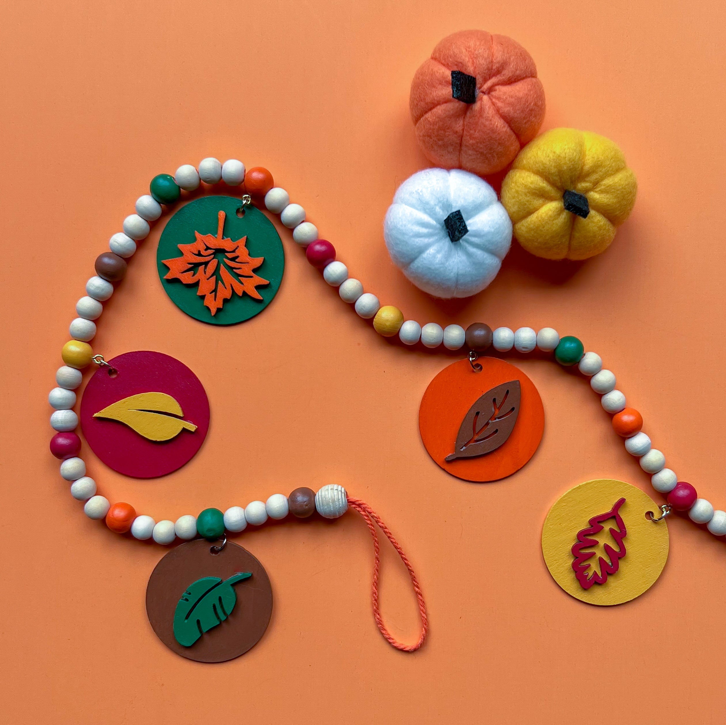 Image shows a wood bead garland with 2" wood circles that feature various colored fall leaves. Circles hang from painted beads with natural wood beads are interspersed. Beads are strung on 2mm orange macrame cord with 3" loops on each end.