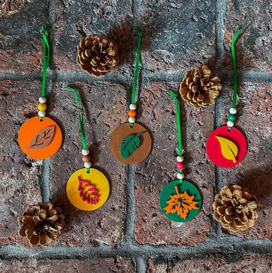 Fall themed ornaments featuring five 2-inch wood circles with wood leaves painted green, red, yellow, orange, or brown. Wood shapes hang from a green ribbon with three wood beads.