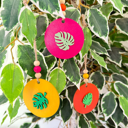 Set of 3 wood, tropical themed ornaments. Ornaments are flat wood circles measuring 2.75 inches; each features a different monstera or palm leaf. Ornaments are painted in magenta, yellow, or orange and include three wood beads strung on jute twine.