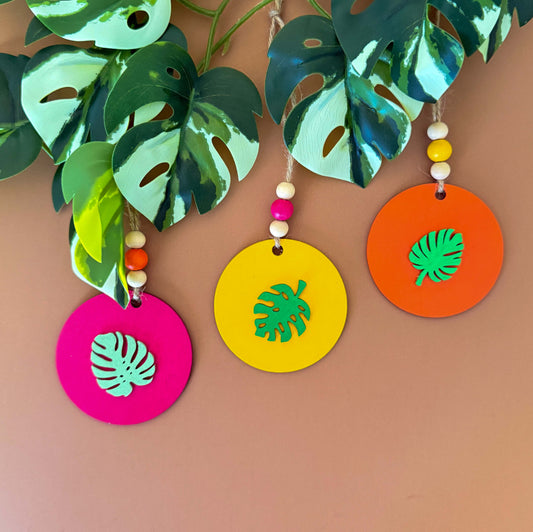 Set of 3 wood, tropical themed ornaments. Ornaments are flat wood circles measuring 2.75 inches; each features a different monstera or palm leaf. Ornaments are painted in magenta, yellow, or orange and include three wood beads strung on jute twine.