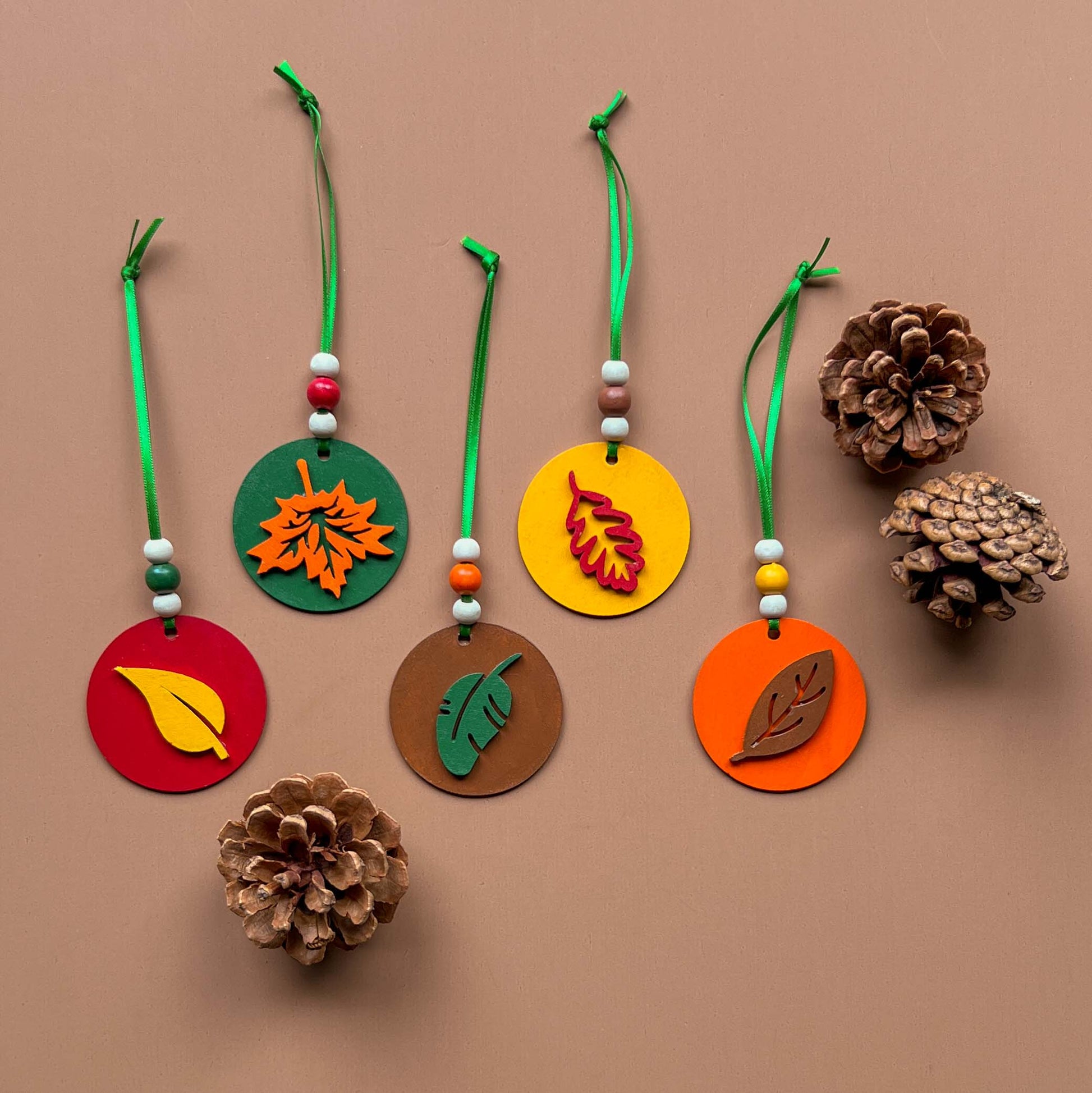 Fall themed ornaments featuring five 2-inch wood circles with wood leaves painted green, red, yellow, orange, or brown. Wood shapes hang from a green ribbon with three wood beads.