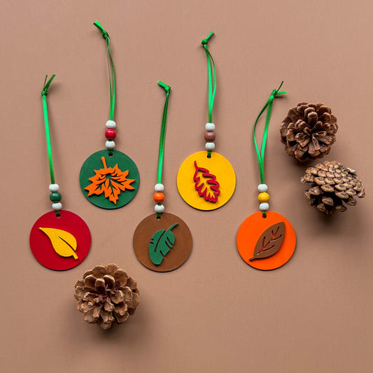 Fall themed ornaments featuring five 2-inch wood circles with wood leaves painted green, red, yellow, orange, or brown. Wood shapes hang from a green ribbon with three wood beads.
