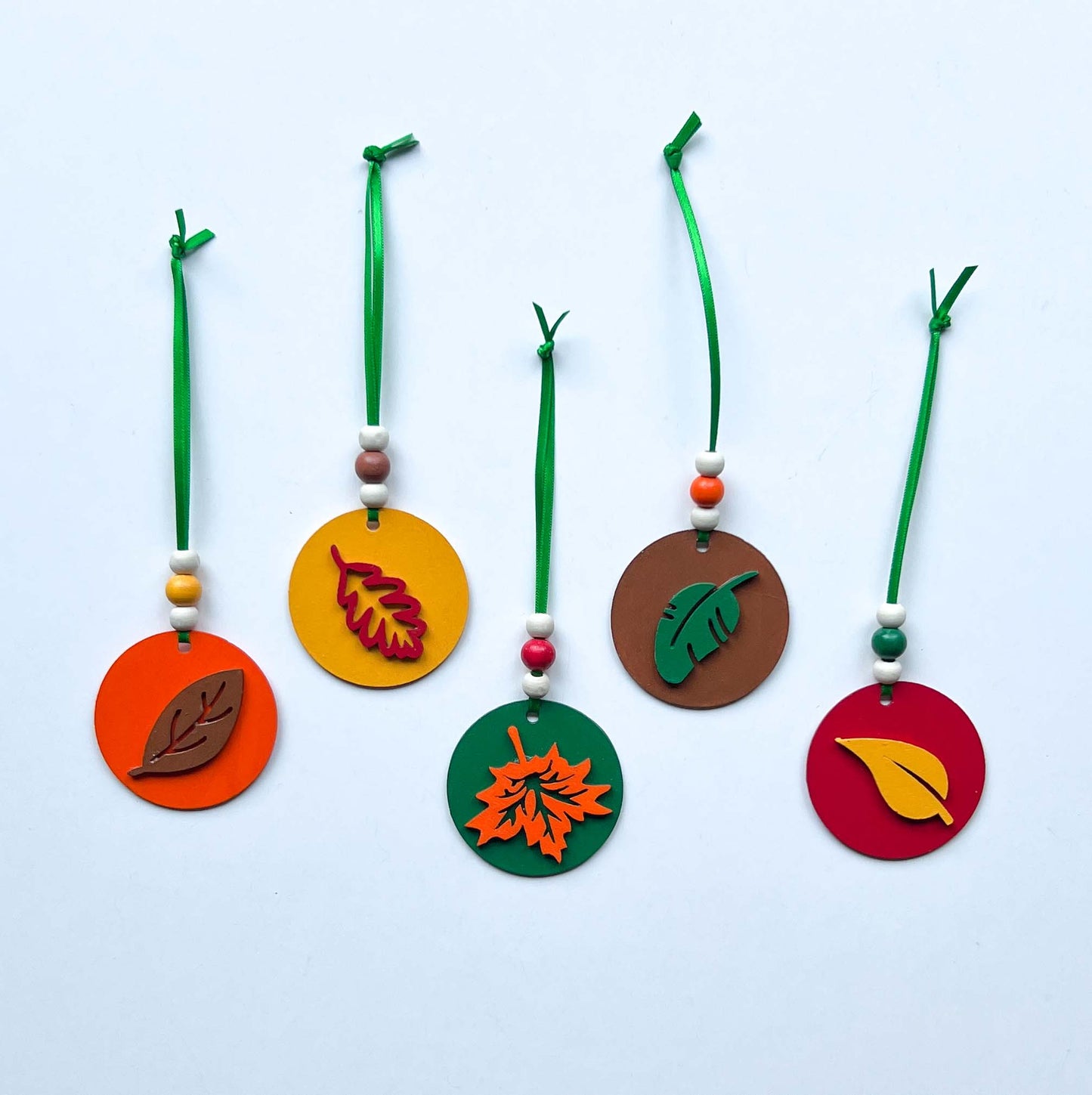 Fall themed ornaments featuring five 2-inch wood circles with wood leaves painted green, red, yellow, orange, or brown. Wood shapes hang from a green ribbon with three wood beads.