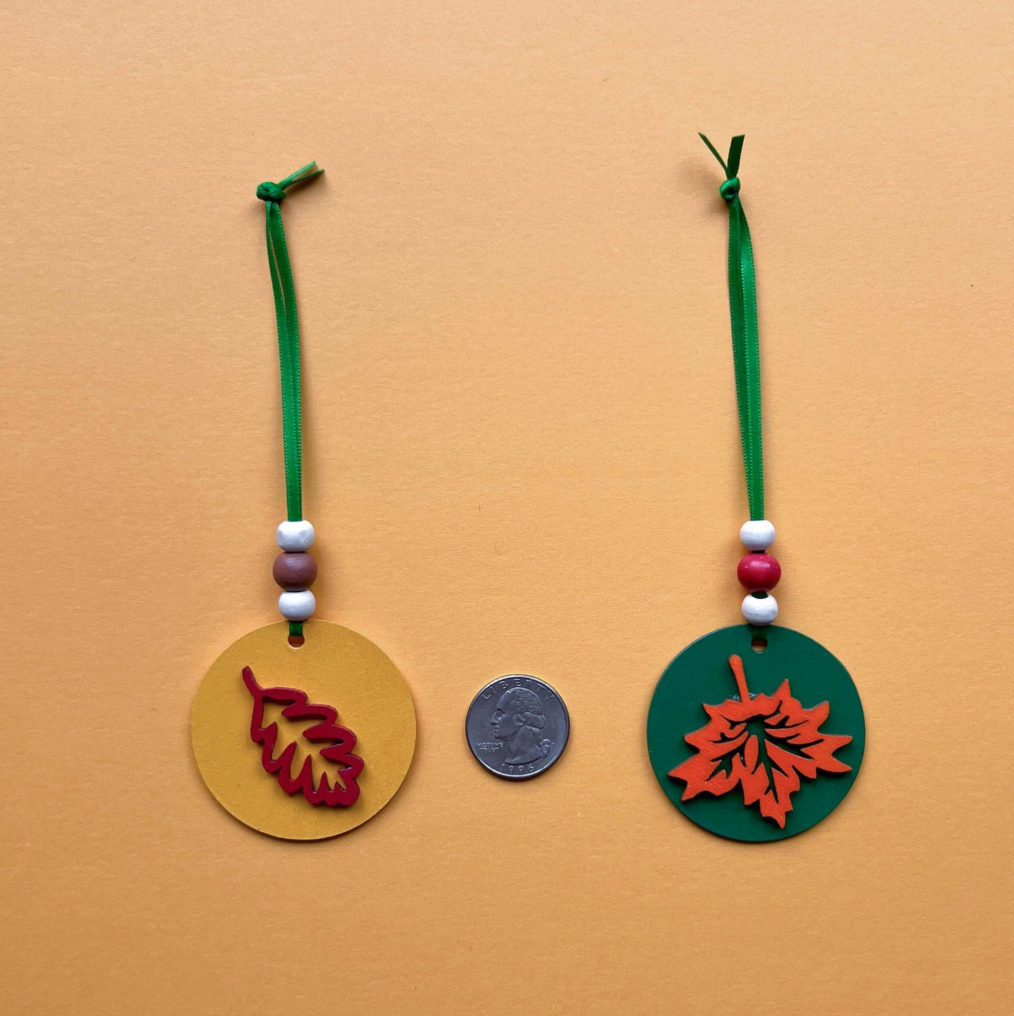 Fall themed ornaments featuring five 2-inch wood circles with wood leaves painted green, red, yellow, orange, or brown. Wood shapes hang from a green ribbon with three wood beads.