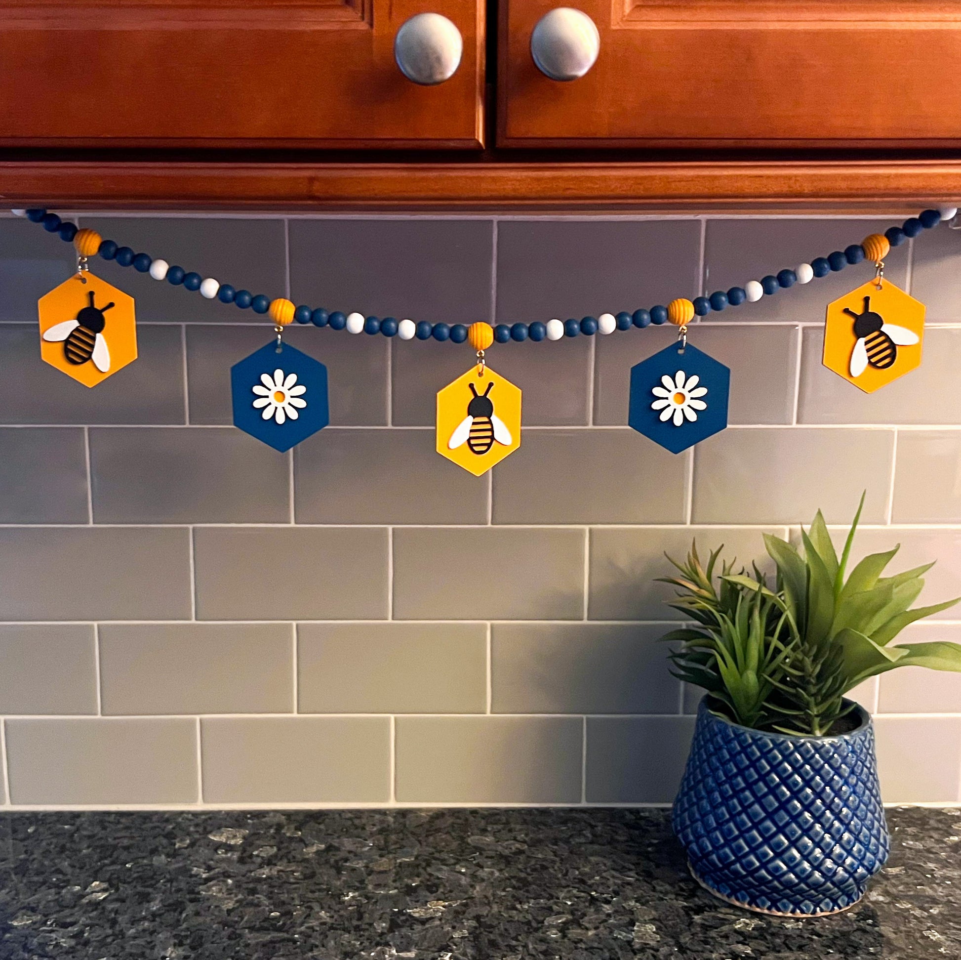 The image shows a wood bead garland featuring wood hexagons with bees and flowers, strung with beads painted deep blue, golden yellow, and white. Hexagons hang from beehive shaped beads. Beads are strung on 2mm blue macrame cord with 3-inch loops on each end.