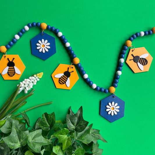 The image shows a wood bead garland featuring wood hexagons with bees and flowers, strung with beads painted deep blue, golden yellow, and white. Hexagons hang from beehive shaped beads. Beads are strung on 2mm blue macrame cord with 3-inch loops on each end.