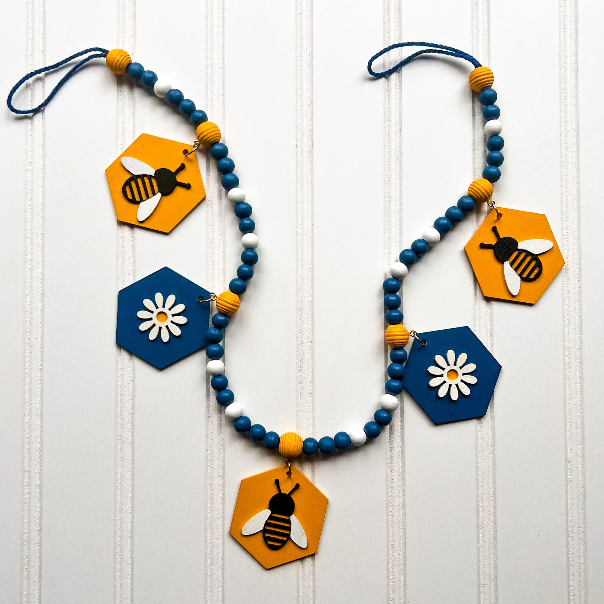 The image shows a wood bead garland featuring wood hexagons with bees and flowers, strung with beads painted deep blue, golden yellow, and white. Hexagons hang from beehive shaped beads. Beads are strung on 2mm blue macrame cord with 3-inch loops on each end.